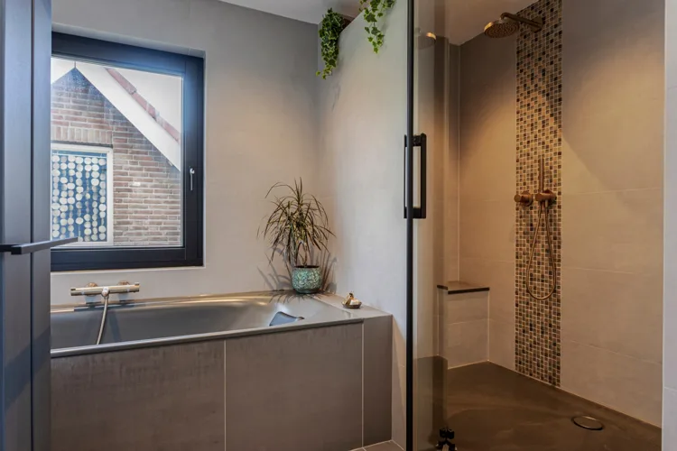 Bad onder een raam naast een inloopdouche met warme tegels.